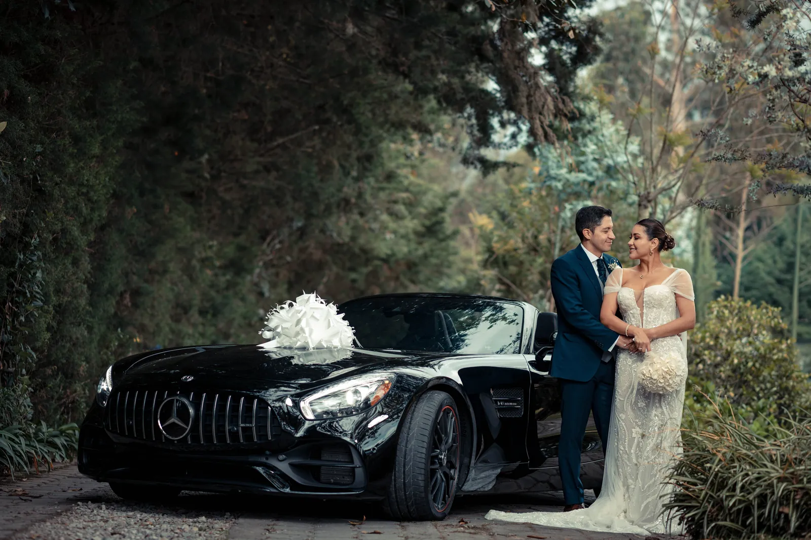 Los novios junto al auto de bodas en un camino arbolado de Villa Fiorenza