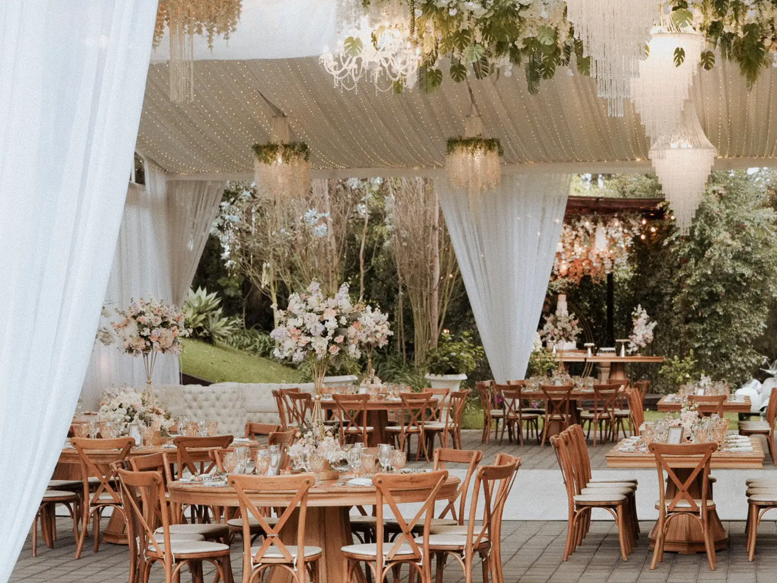 Salon de recepcion montado con mesas redondas, sillas de madera y lamparas colgantes entre flores en Villa Fiorenza, quinta para bodas en Puembo, Quito